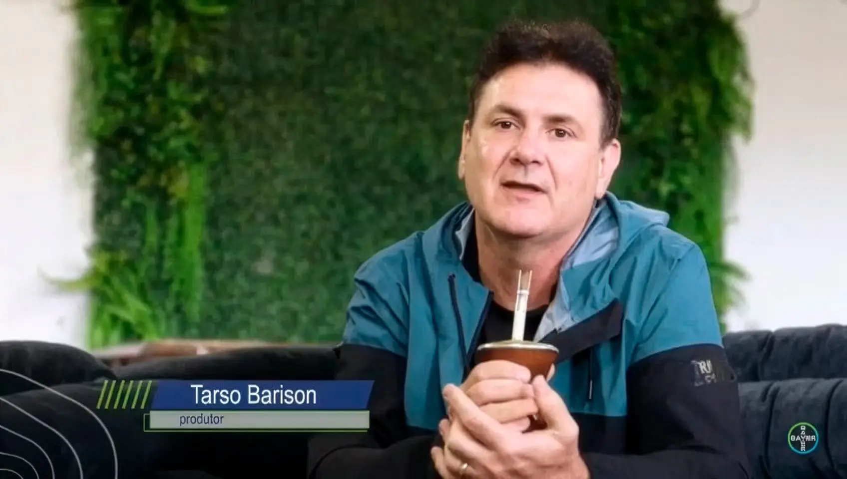 Tarso Barison, identificado na tela como produtor, sentado num sofá escuro com uma cuia de chimarrão nas mãos; ao fundo, uma parede de folhagem verde